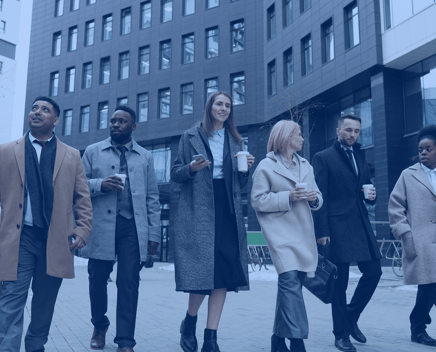 Groupe de professionnels marchant ensemble dans un quartier d’affaires moderne, habillés en tenue formelle avec des manteaux d’hiver et des gobelets à la main, illustrant la diversité, la collaboration et la convivialité en entreprise.