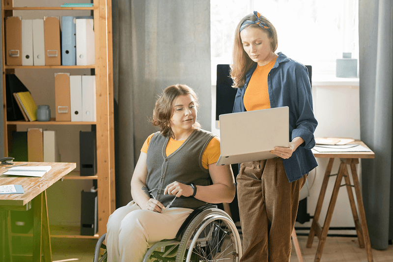 Une femme en fauteuil roulant discute avec une collègue qui lui présente des informations sur un ordinateur portable. Scène dans un bureau moderne et lumineux, illustrant l’inclusion des personnes en situation de handicap au travail.