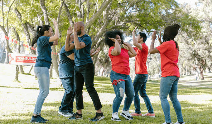 Groupe de collègues en pleine activité ludique en extérieur, se tapant dans les mains par binômes sous une bannière “Teambuilding Event”, dans un parc ensoleillé entouré d’arbres.