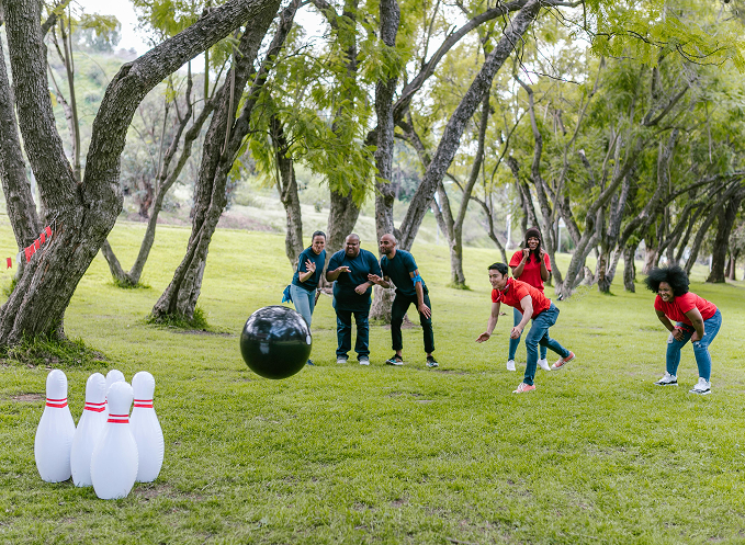 Groupe de collègues lançant une grosse boule noire vers des quilles blanches sur une pelouse entourée d’arbres, lors d’un jeu collectif en extérieur.