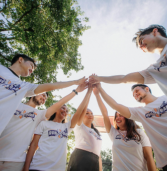 Groupe de jeunes adultes en t-shirts blancs, formant un cercle et levant les mains en signe d’unité lors d’un événement d’équipe en plein air, sous un ciel dégagé.