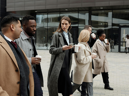 Groupe de professionnels marchant ensemble devant un bâtiment moderne, habillés en tenue de bureau avec des manteaux d’hiver et des gobelets à la main, discutant en chemin.