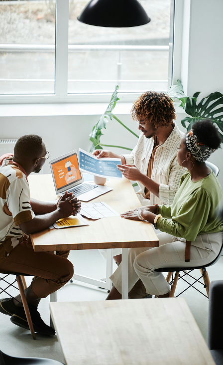 Trois collègues en pleine discussion autour d’une table dans un bureau moderne, analysant des supports visuels et un ordinateur affichant une présentation colorée, dans un cadre lumineux avec des plantes d’intérieur.