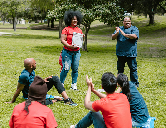 Femme souriante debout avec un certificat en main, félicitée par ses collègues assis dans l’herbe pendant une activité de team building en plein air.