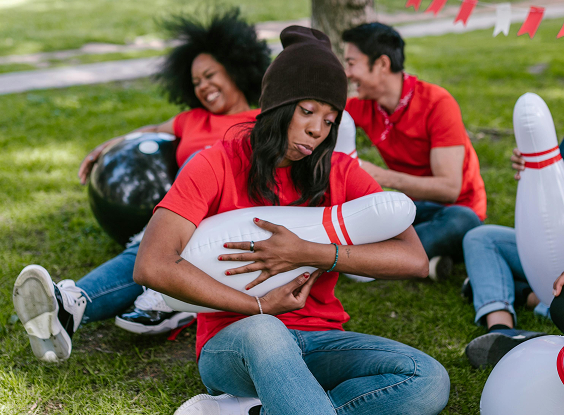 Jeune femme en t-shirt rouge assise dans l’herbe, tenant une quille de bowling gonflable, entourée de collègues riant dans une ambiance décontractée.